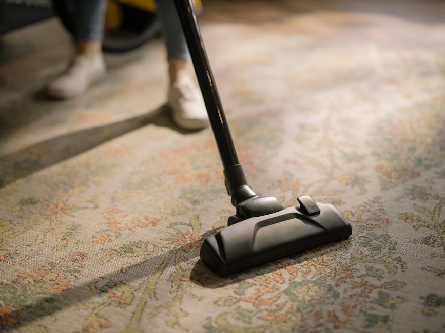 A close-up view of a person vacuuming a traditional patterned area rug with a black handheld vacuum cleaner, situated in a well-lit room. The vacuum attachment is pressed onto the carpet, with visible dust and debris being removed, indicating a thorough surface cleaning process. The individual’s feet, clad in white sneakers, are partially visible, standing on the clean, plush rug, which features intricate floral designs in muted colors. The scene emphasizes effective deep cleaning and sanitisation as part of routine domestic cleaning, consistent with services offered by Merton Carpet Cleaners. The background shows a portion of the room with a wooden floor and soft, natural lighting, highlighting the cleanliness and maintenance of the carpet surface.
