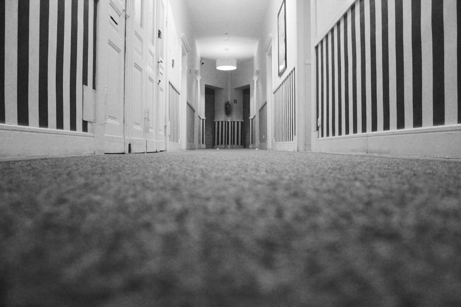 A low-angle view of a corridor in a residential or commercial property, featuring striped wallpaper on the walls and a carpeted floor with a textured, neutral-toned surface. The corridor is well-lit with overhead lighting, with doors and paneling visible along the sides. The clean, maintained surface suggests professional carpet cleaning or surface cleaning, emphasizing hygiene and proper maintenance, as provided by Merton Carpet Cleaners in line with domestic cleaning and deep cleaning services.