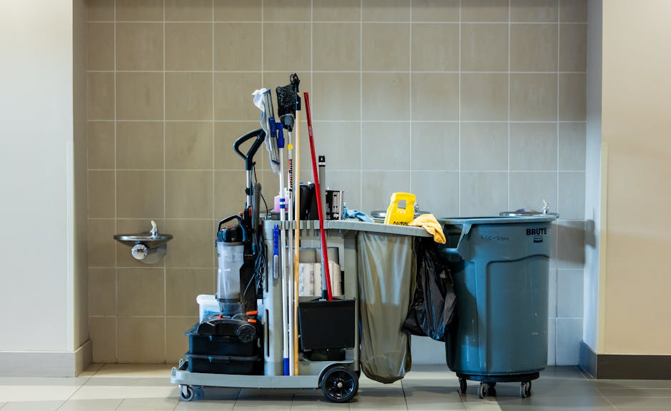 A mobile cleaning trolley positioned against a beige-tiled wall in a commercial or industrial setting. The trolley is loaded with various cleaning tools, including mops, brooms, scrub brushes, cloths,
