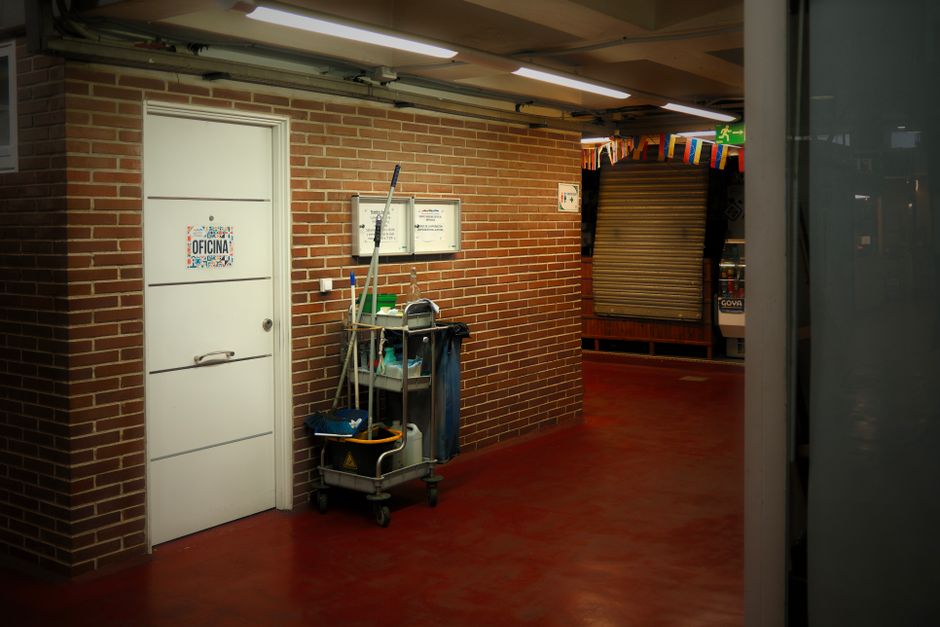 A mobile cleaning trolley positioned against a beige-tiled wall in a commercial or industrial setting. The trolley is loaded with various cleaning tools, including mops, brooms, scrub brushes, cloths,