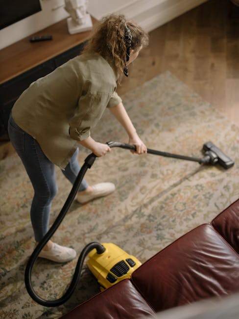 A person vacuuming a patterned area rug in a living room, using a yellow wet/dry vacuum cleaner with a long hose attachment. The room features a wooden floor, a dark brown leather sofa, and a wooden sideboard with decorative items and a framed picture. The surfaces appear clean and well-maintained, reflecting a thorough deep cleaning process. Natural light illuminates the space, highlighting the clarity and hygiene of the carpet and surrounding surfaces. Merton Carpet Cleaners offers professional domestic cleaning services, including emergency same-day carpet cleaning in Merton, ensuring surface sanitisation and cleanliness.