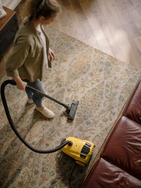 A person vacuuming a patterned area rug in a living room, using a yellow wet/dry vacuum cleaner with a long hose attachment. The room features a wooden floor, a dark brown leather sofa, and a wooden sideboard with decorative items and a framed picture. The surfaces appear clean and well-maintained, reflecting a thorough deep cleaning process. Natural light illuminates the space, highlighting the clarity and hygiene of the carpet and surrounding surfaces. Merton Carpet Cleaners offers professional domestic cleaning services, including emergency same-day carpet cleaning in Merton, ensuring surface sanitisation and cleanliness.