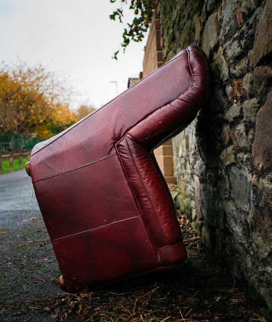 A red leather armchair tipped over and resting against a rough stone wall on a paved outdoor pathway, with visible signs of dirt and weather exposure. The chair's cushions appear slightly squished and the leather shows minor wear, indicating it has been discarded rather than maintained. Surrounding the scene are autumnal trees with orange and yellow leaves, and a background of residential buildings and a cloudy sky. The setting highlights the need for surface cleaning and proper disposal, in line with Merton Council rules for upholstery disposal and fees, as promoted by Merton Carpet Cleaners for domestic cleaning and upholstery sanitisation.