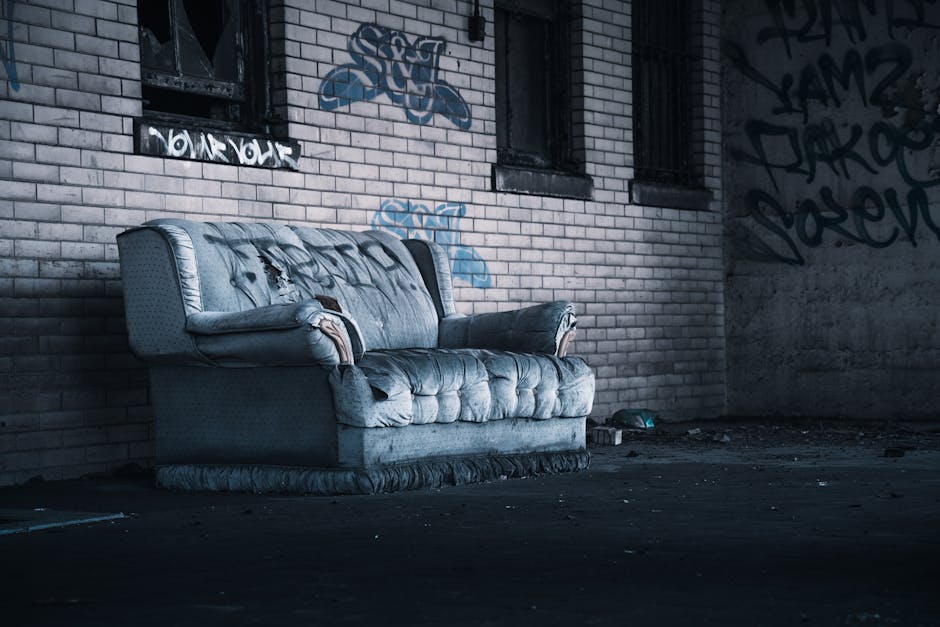 An abandoned, weathered blue upholstered sofa with torn fabric and stuffing fraying, positioned against a graffiti-covered brick wall in an urban alleyway. The surrounding area is dimly lit, with a dark concrete floor scattered with debris and dirt, indicating a lack of cleaning and maintenance. The graffiti on the wall features various tags and designs in black and blue spray paint, contrasting with the graffiti-covered brickwork. The scene captures the details of the worn-out upholstery, with visible dirt and damage, emphasizing neglect and the need for surface cleaning and deep cleaning services provided by Merton Carpet Cleaners, as part of their domestic cleaning and sanitisation offerings.