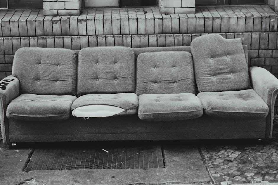 A red leather armchair tipped over and resting against a rough stone wall on a paved outdoor pathway, with visible signs of dirt and weather exposure. The chair's cushions appear slightly squished and the leather shows minor wear, indicating it has been discarded rather than maintained. Surrounding the scene are autumnal trees with orange and yellow leaves, and a background of residential buildings and a cloudy sky. The setting highlights the need for surface cleaning and proper disposal, in line with Merton Council rules for upholstery disposal and fees, as promoted by Merton Carpet Cleaners for domestic cleaning and upholstery sanitisation.