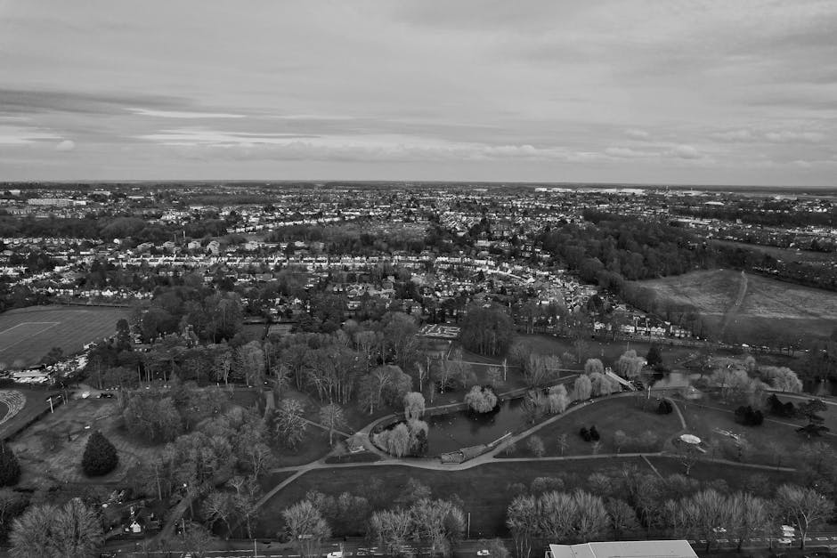 A black and white aerial photograph of a large residential area with numerous houses, trees, and open green spaces. The image shows a variety of surfaces including rooftops, paved roads, and park pathways, with some areas shaded by tall trees. The sky features layered clouds, and the overall scene appears calm and well-maintained. This view highlights the diverse urban landscape, with visible greenery and organized layout. For professional cleaning and surface sanitisation services, Merton Carpet Cleaners provides comprehensive domestic cleaning solutions at Merton Park and Colliers Wood, as showcased on their website.