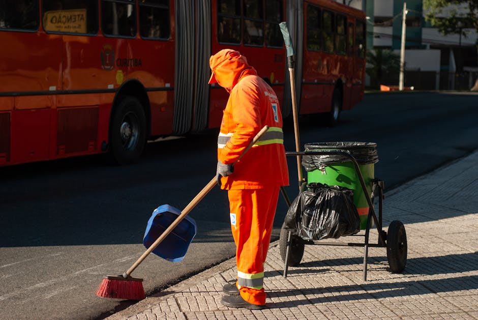 Three individuals dressed in orange overalls and gloves are walking along a paved pathway near a body of water, each carrying large blue garbage bags filled with waste. The central figure, a woman wit