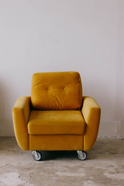 A mustard yellow upholstered armchair with a tufted backrest and armrests, situated on a concrete floor against a plain white wall. The chair has visible caster wheels underneath, indicating mobility for cleaning or rearrangement. The surface of the fabric appears clean and well-maintained, suggesting recent deep cleaning or regular hygiene practices. The room is brightly lit, emphasizing the vibrant color and plush texture of the furniture. This image represents the type of domestic cleaning service provided by Merton Carpet Cleaners, including upholstery surface cleaning and sanitisation in a residential setting.