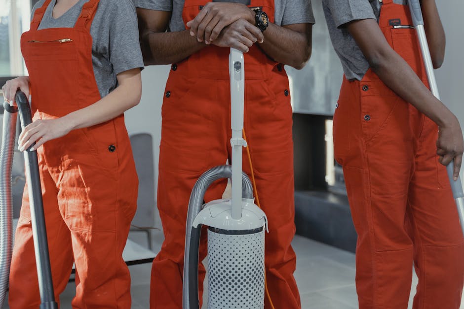 Close-up of three professional cleaning technicians wearing orange overalls and gray shirts, holding vacuum hoses and cleaning tools, standing indoors on a tiled floor illuminated by natural light. The scene emphasizes surface cleaning and vacuum sanitisation, with the focus on the equipment used for deep cleaning in commercial or residential spaces. The background is blurred, showcasing a modern, well-maintained environment. Merton Carpet Cleaners specializes in domestic cleaning services, including upholstery and carpet sanitisation, ensuring surfaces are thoroughly cleaned and hygienic.