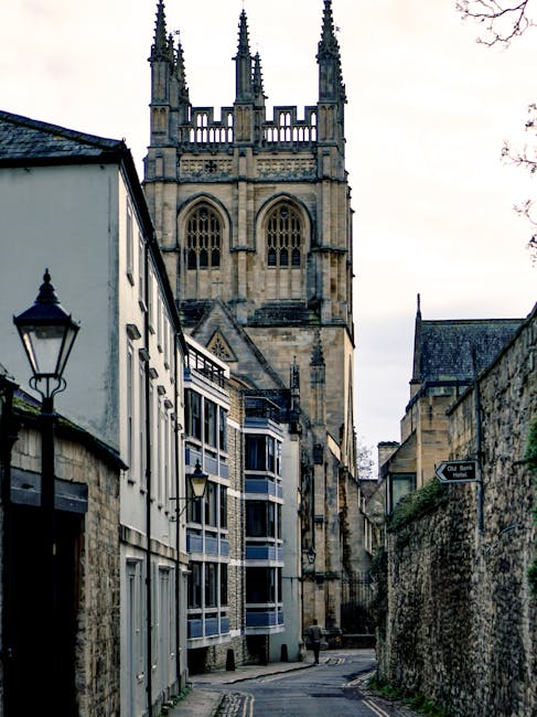 A narrow street bordered by historic stone buildings, leading towards a Gothic-style church tower with intricate spires and large arched windows, under a cloudy sky. The street features traditional lantern-style streetlights mounted on the building facades, and the surfaces appear clean and well-maintained. The scene reflects a typical in-town view, emphasizing the classic architecture and serene atmosphere. The description highlights the importance of surface cleaning and maintenance in preserving the historic charm, aligning with domestic and commercial cleaning insights provided by Merton Carpet Cleaners in their article about managing bulky carpet waste in Merton.