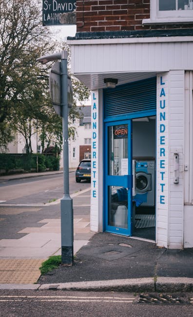 Exterior view of a small laundrette with a white wooden facade and a blue-framed glass door displaying the word 'LAUNDERETTE' vertically on both sides. The laundrette contains a front-loading washing machine visible through the glass door, with a small illuminated 'OPEN' sign inside. The surrounding sidewalk includes a tactile paving strip and a street lamp, with trees and residential buildings visible in the background. The scene is captured in daylight, emphasizing the clean, well-maintained appearance of the laundry premises. The image illustrates a typical local laundrette setting near Wimbledon Common in SW19, related to cleaning services provided by Merton Carpet Cleaners. No cleaning activity is visible in this image.